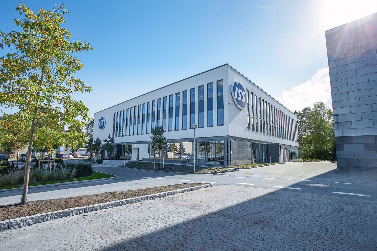 Ein modernes, rechteckiges Bürogebäude mit großen Fenstern und dem Logo "ISS" an der Fassade, umgeben von Bäumen und einem gepflasterten Vorplatz, unter einem klaren, blauen Himmel.