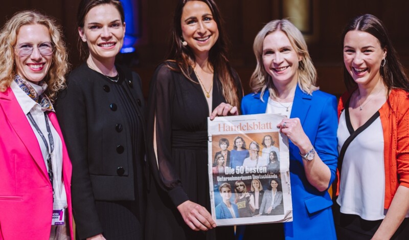 Fünf Frauen stehen lächelnd zusammen und halten eine Ausgabe des Handelsblatt hoch, die einen Artikel über die "50 besten Unternehmerinnen Deutschlands" zeigt. Sie sind in farbenfrohe Kleidung gekleidet, und das Bild scheint bei einer Veranstaltung oder Preisverleihung aufgenommen worden zu sein.