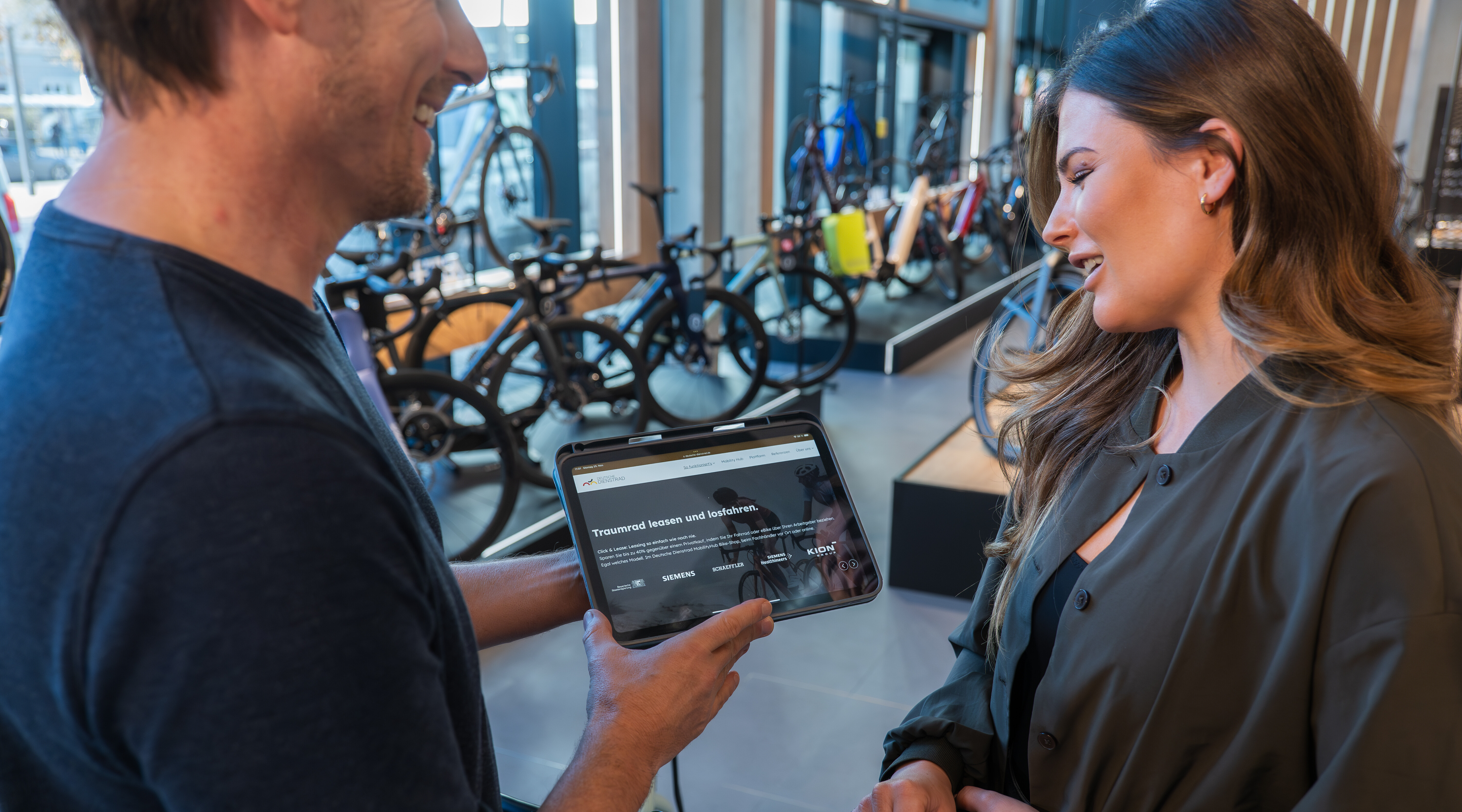 Ein Mann zeigt einer Frau in einem Fahrradgeschäft ein Tablet mit einer Leasing-Webseite, während im Hintergrund viele Fahrräder ausgestellt sind.