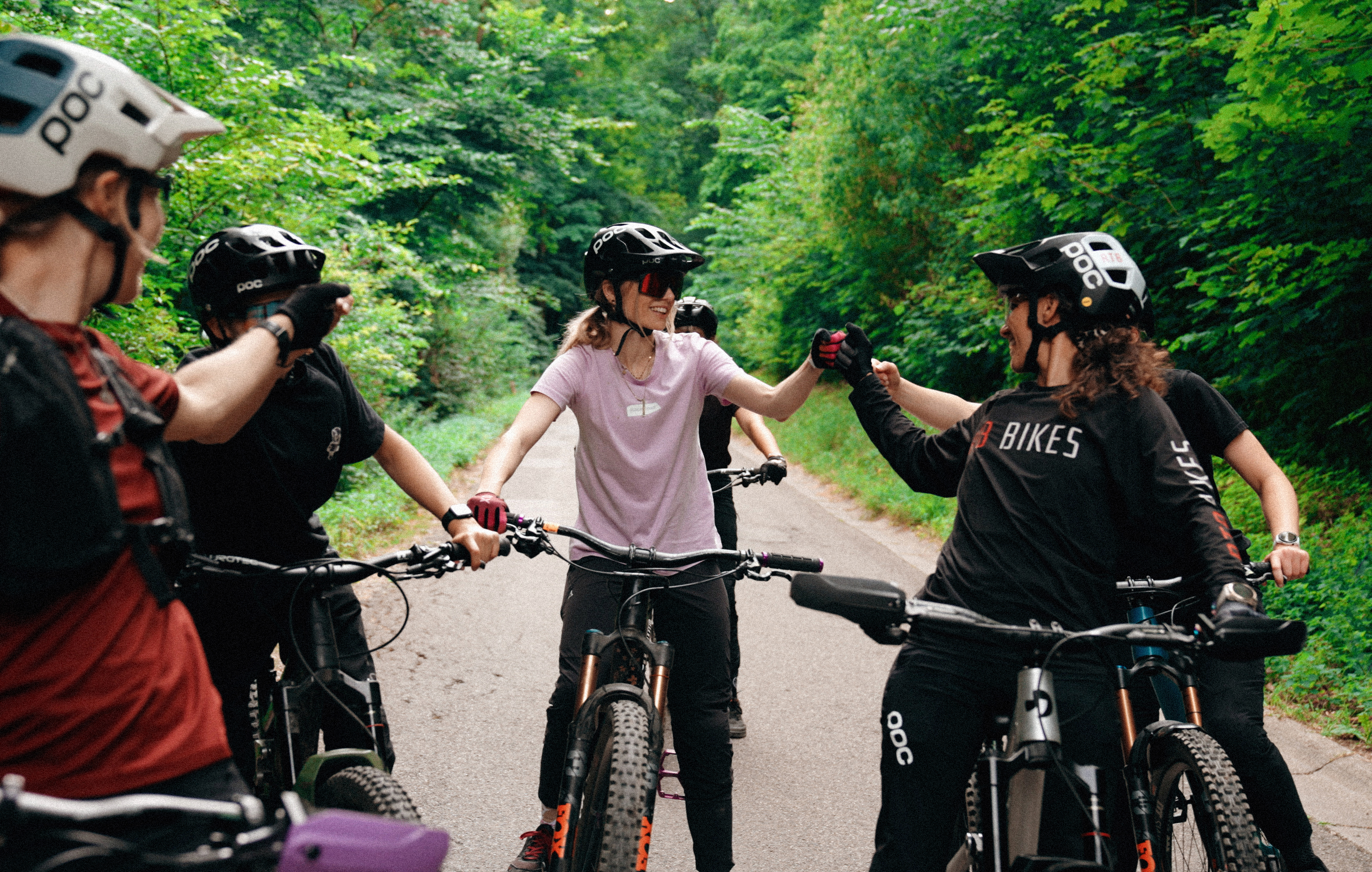 Eine Gruppe von Radfahrerinnen auf einer Waldstraße gibt sich lachend Fistbumps.