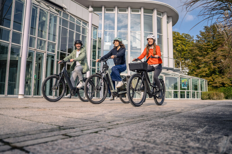 Drei Personen fahren mit E-Bikes lächelnd vor einem modernen, verglasten Gebäude.
