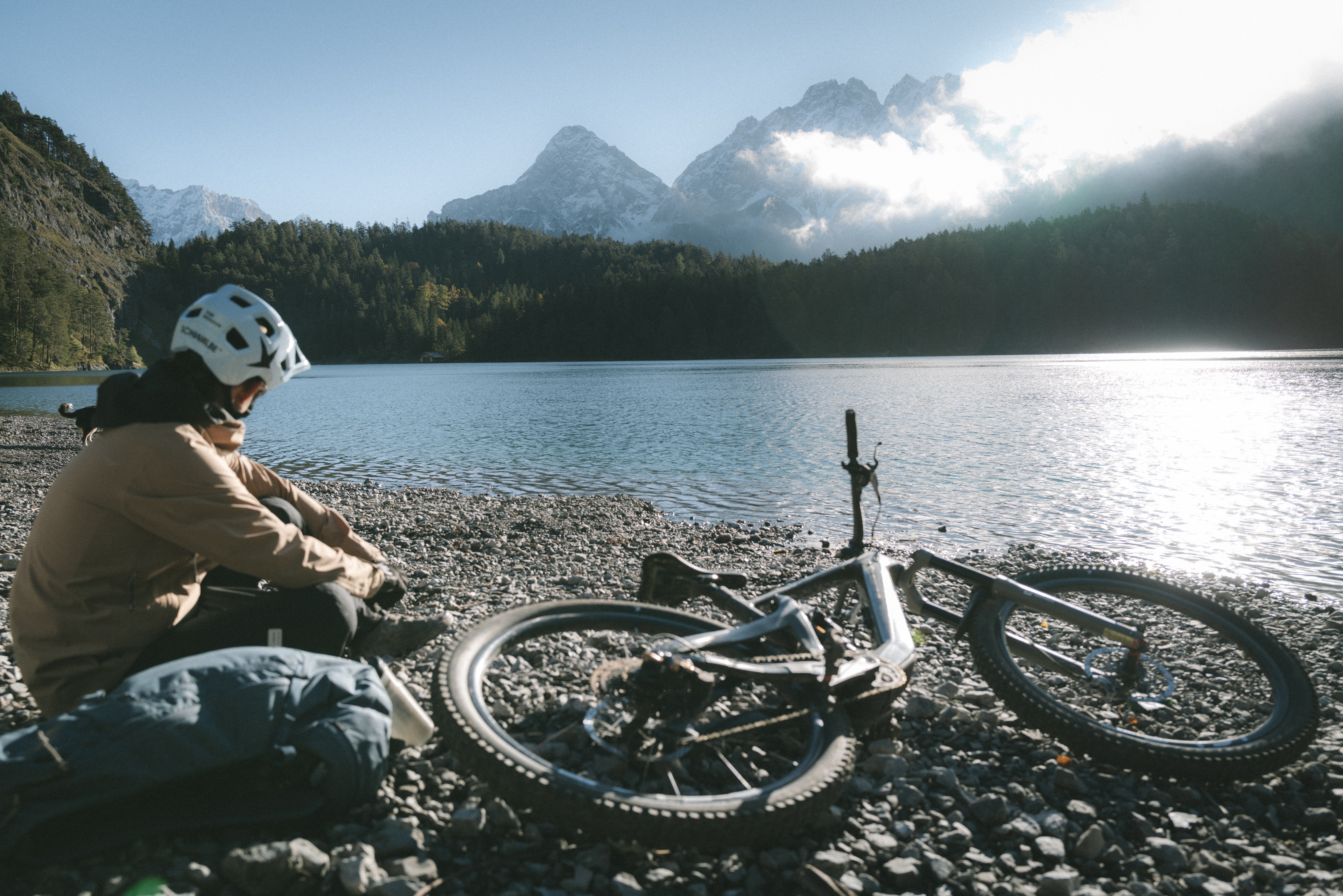Eine Person mit Fahrradhelm sitzt am Ufer eines Bergsees neben einem Mountainbike, im Hintergrund schneebedeckte Gipfel.