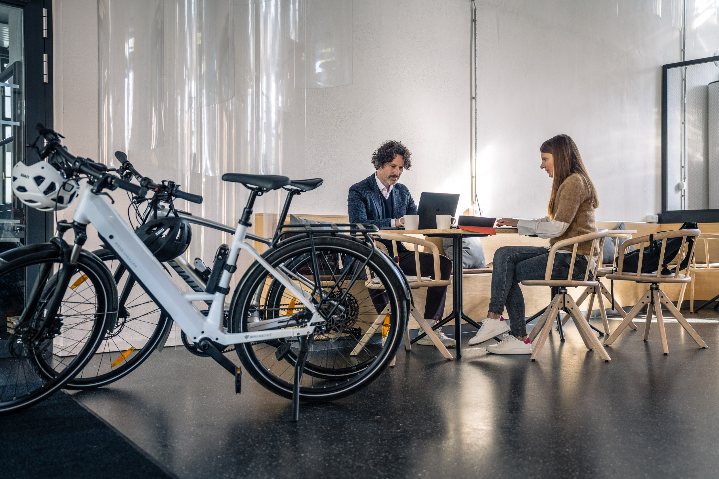 Zwei Personen arbeiten an einem Tisch mit Laptops, während im Vordergrund zwei Fahrräder mit Helmen stehen.