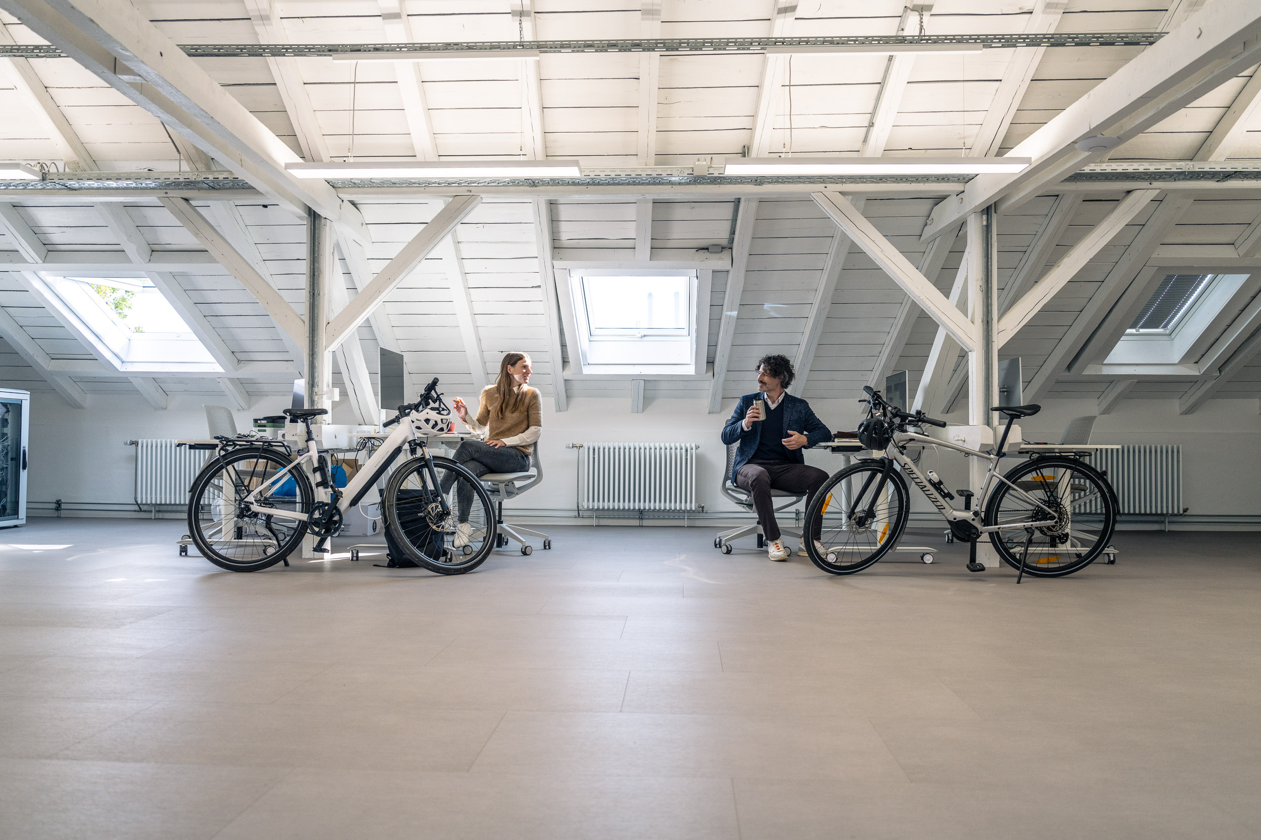 Zwei Personen unterhalten sich in einem hellen Dachgeschossbüro, jeweils neben einem E-Bike sitzend.
