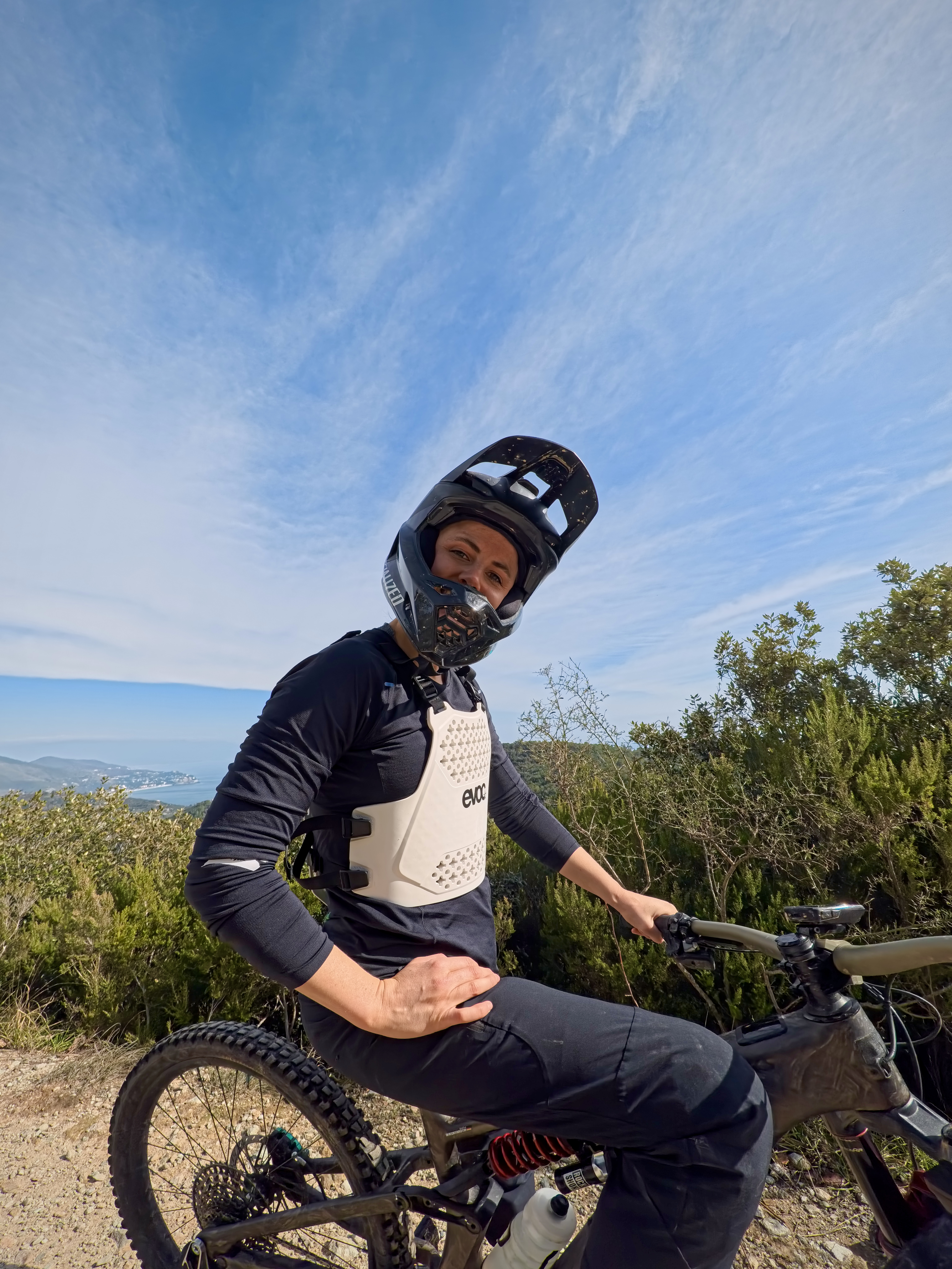 Mountainbiker mit Fullface-Helm und Brustprotektor sitzt auf einem Mountainbike und lächelt in die Kamera; im Hintergrund sind Büsche, Hügel und ein Blick aufs Meer unter blauem Himmel.