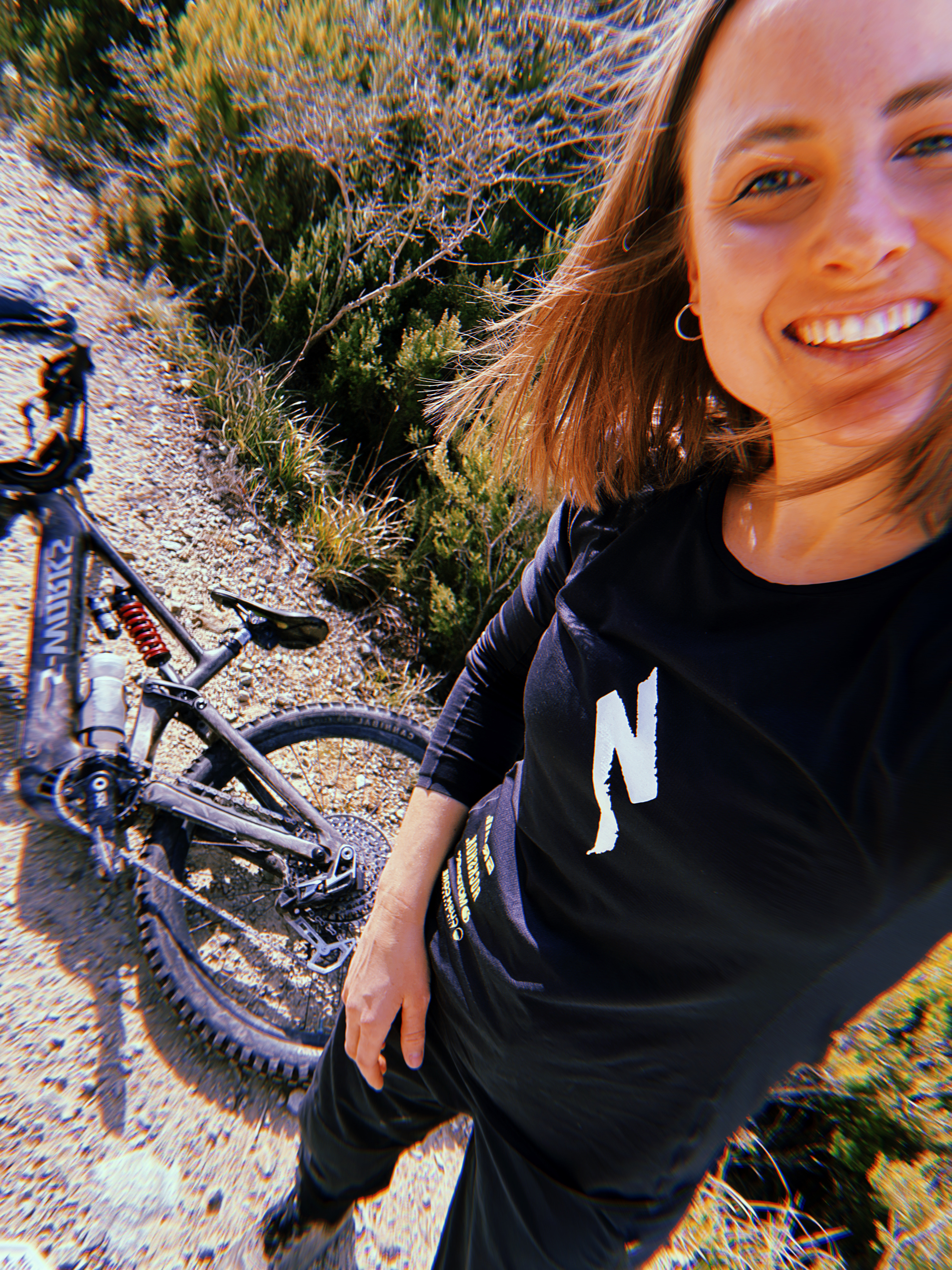 Selfie einer lächelnden Mountainbikerin auf einem sonnigen Schotterweg; neben ihr liegt ein Mountainbike am Boden, im Hintergrund niedrige Büsche und trockenes Gelände.