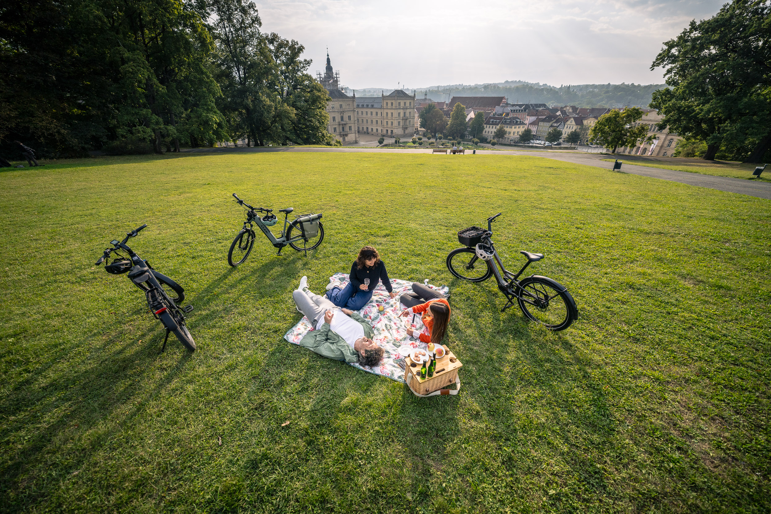 Drei Personen machen ein Picknick auf einer Wiese mit Blick auf eine Altstadt, umgeben von E-Bikes.