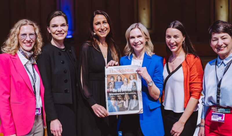 Sechs Frauen lächeln in die Kamera, eine von ihnen hält eine Ausgabe der Zeitung "Handelsblatt", die einen Artikel über die "50 besten Unternehmerinnen Deutschlands" zeigt.