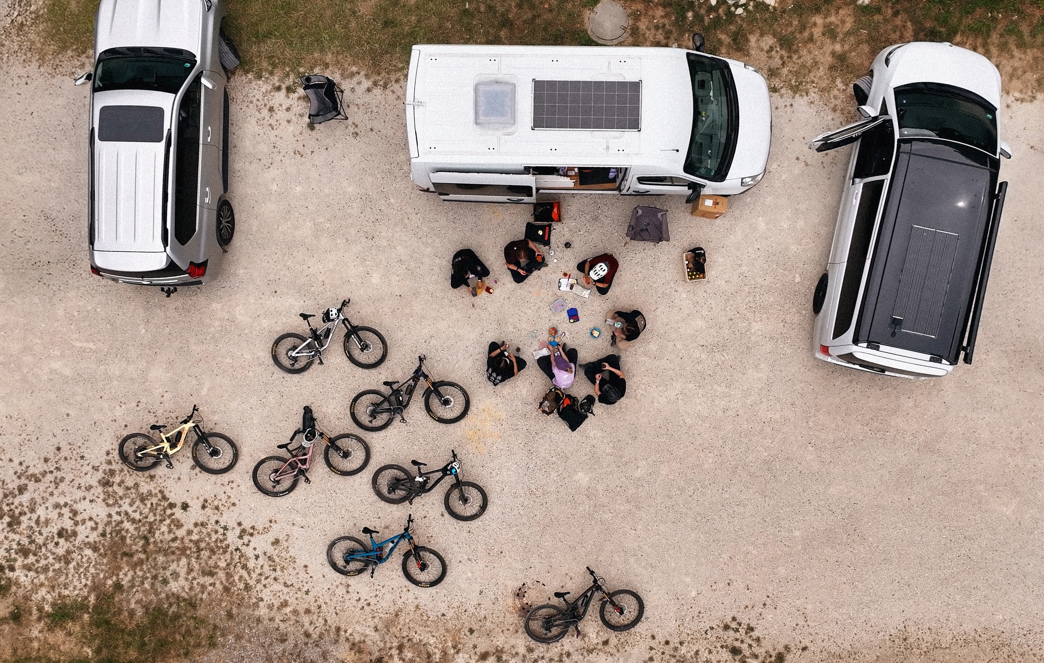 Eine Gruppe von Radfahrerinnen sitzt zwischen Vans und Mountainbikes auf einem Schotterplatz und macht eine Pause.