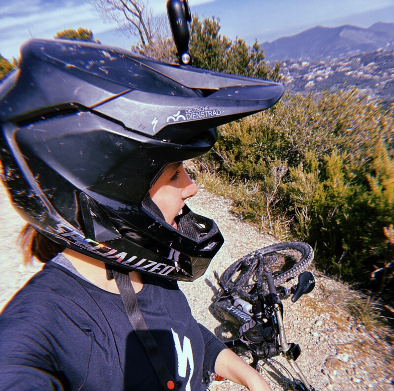 Selfie einer Mountainbikerin mit Fullface-Helm auf einem Schotterweg, das Fahrrad liegt im Hintergrund vor einer Berglandschaft.