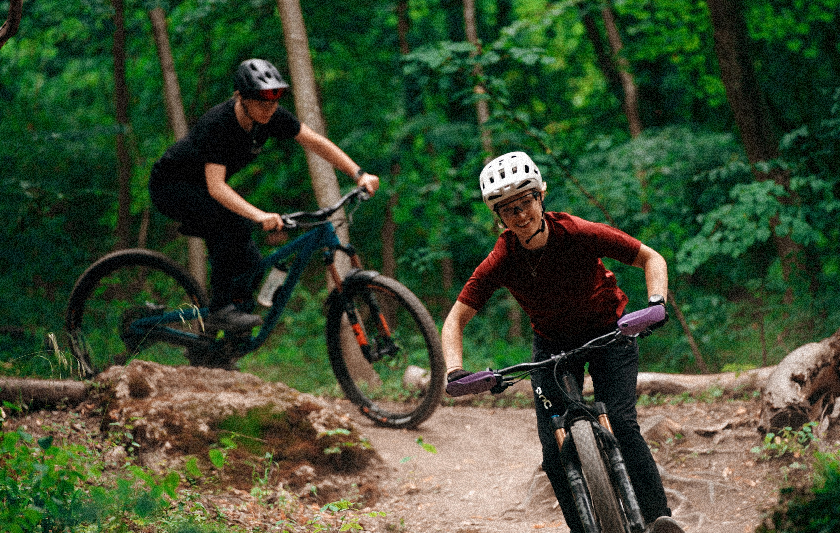 Zwei Radfahrerinnen fahren mit Mountainbikes einen wurzeligen Trail im Wald hinunter.