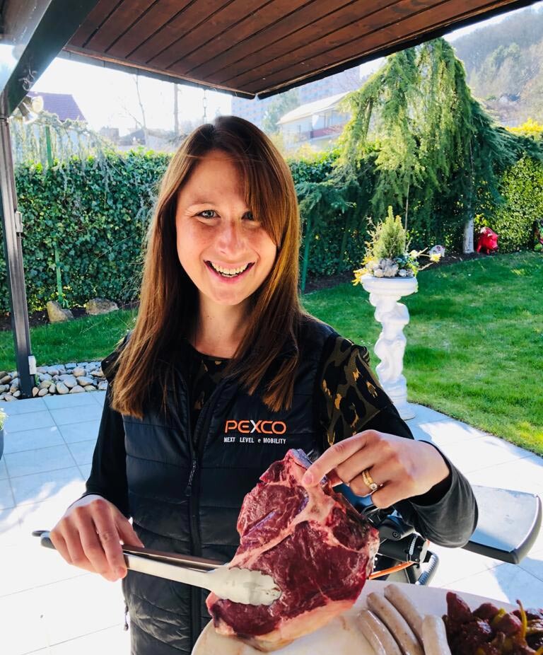 Eine Frau steht draußen unter einem überdachten Bereich und hält ein großes Stück rohes Fleisch mit einer Zange. Sie lächelt in die Kamera und trägt eine schwarze Weste mit dem Aufdruck "Pexco - Next Level Mobility". Im Hintergrund ist ein gepflegter Garten mit Rasen, Büschen und einem dekorativen Pflanzgefäß zu sehen.#