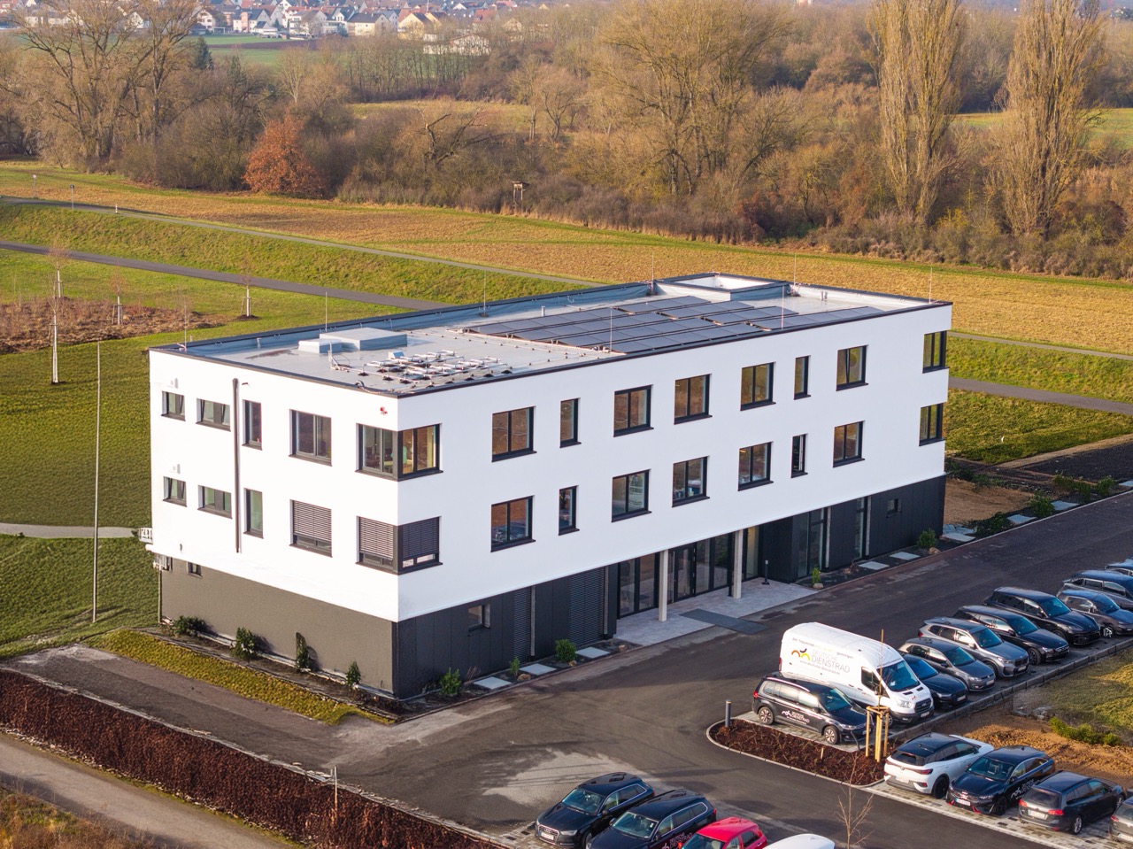 Luftaufnahme eines modernen, dreistöckigen Bürogebäudes mit Solaranlage auf dem Dach, neben einem Parkplatz und umgeben von Feldern und Bäumen.