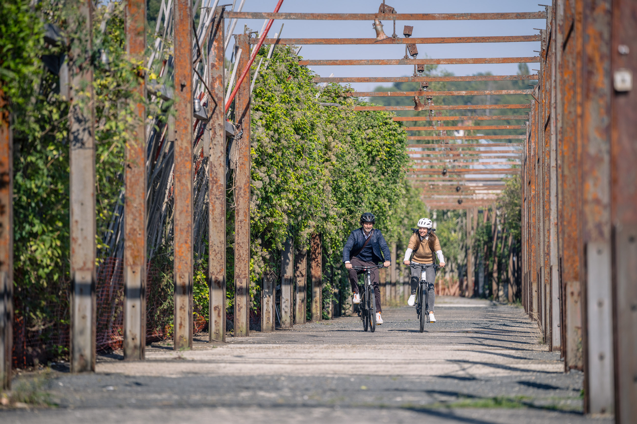 Zwei Personen fahren nebeneinander mit dem Fahrrad durch einen von Pflanzen und rostigen Metallträgern gesäumten Weg.
