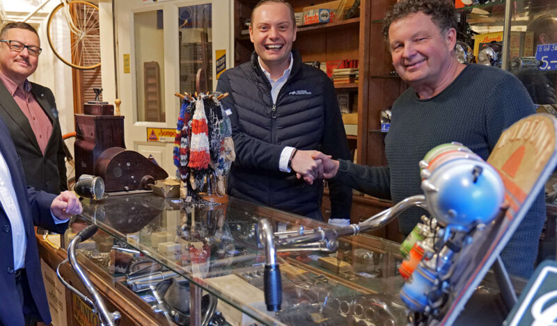 Zwei Männer stehen hinter einer Glasvitrine in einem Geschäft und schütteln sich die Hand. Die Umgebung ist gefüllt mit Vintage-Gegenständen und Fahrradteilen. Beide Männer lächeln in die Kamera.