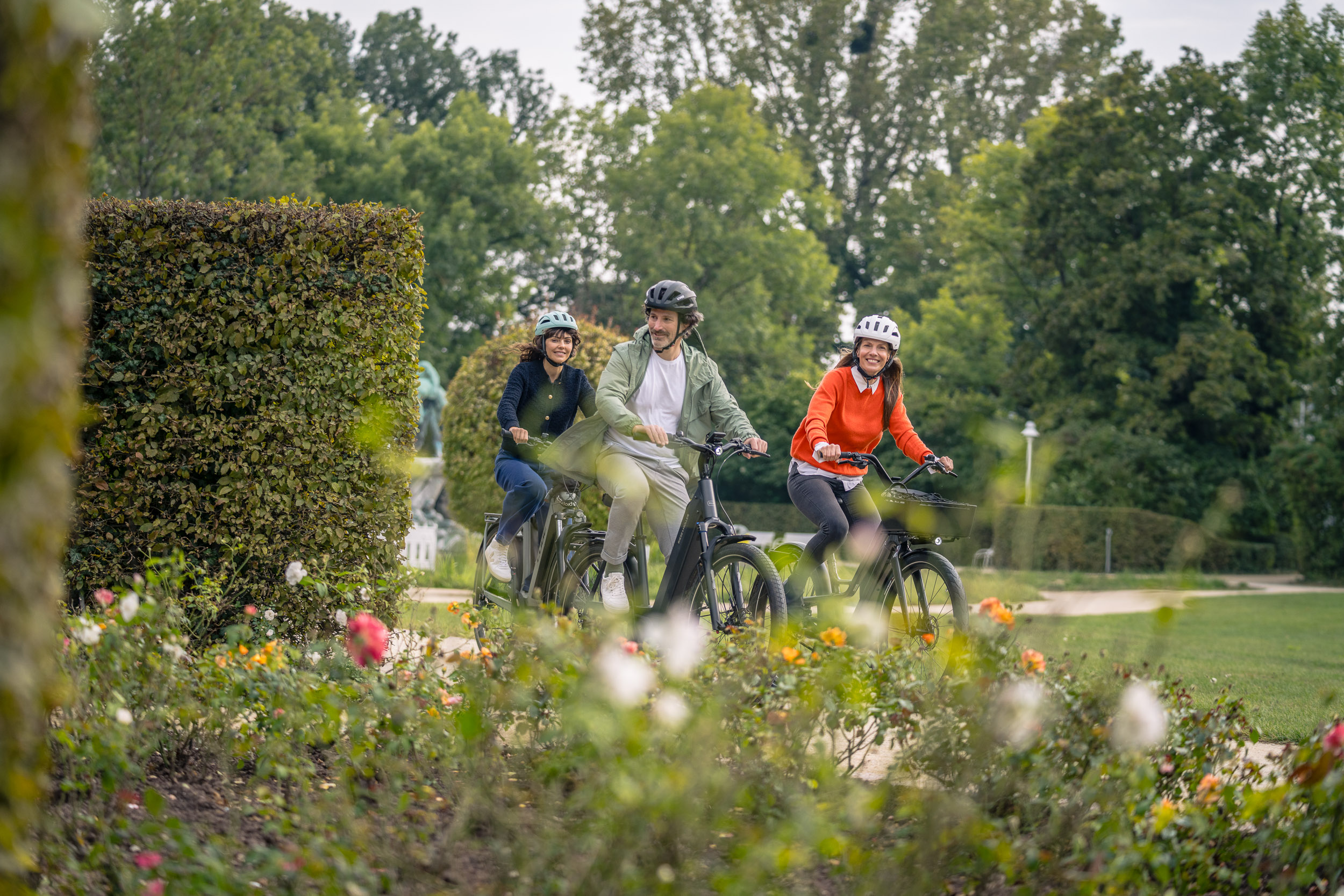 Drei Personen fahren lachend mit dem Fahrrad durch eine gepflegte Parkanlage mit blühenden Rosen.