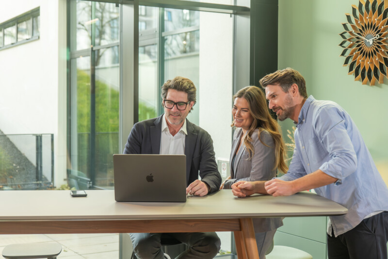 Drei Personen stehen an einem Tisch und schauen gemeinsam auf einen Laptop, während sie lächeln.