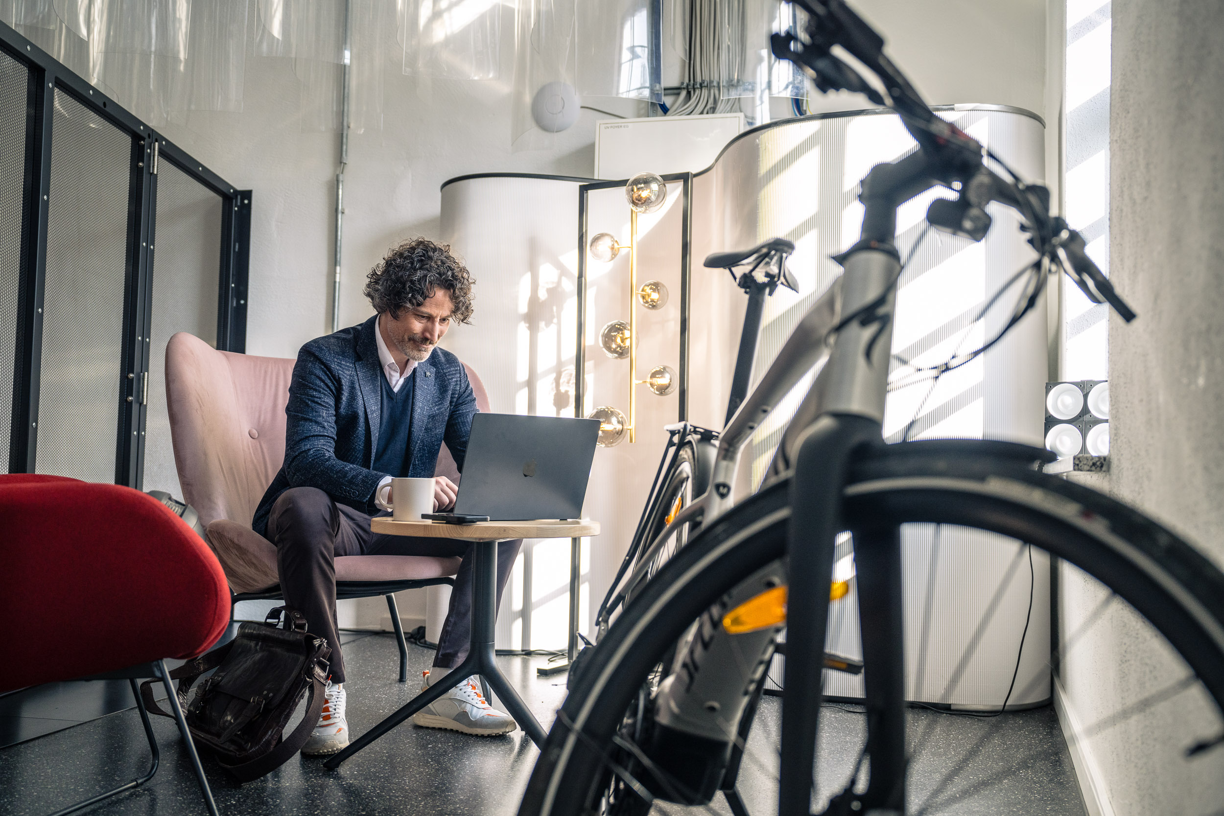 Ein Mann arbeitet mit einem Laptop in einem modern eingerichteten Raum, während ein Fahrrad im Vordergrund steht.
