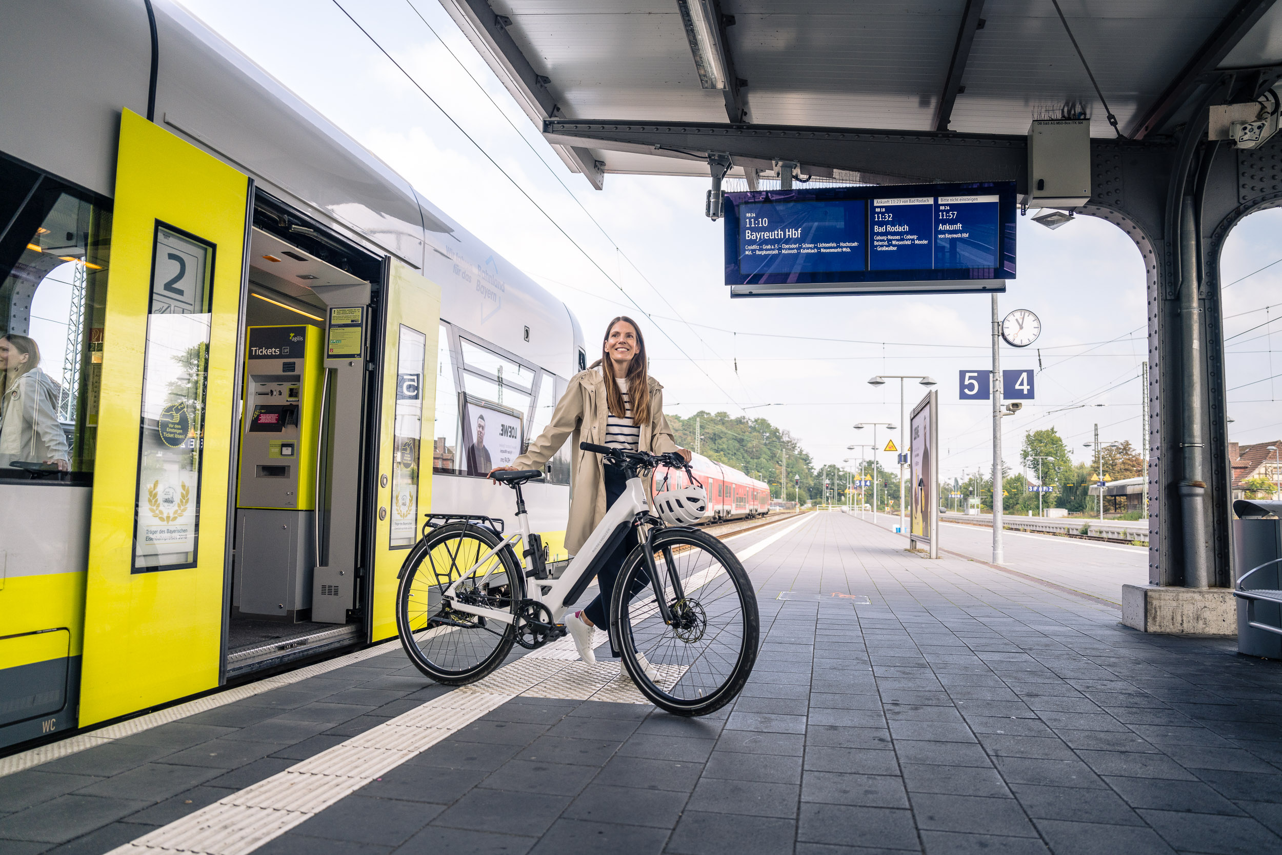 Eine Frau steht mit einem E-Bike auf dem Bahnsteig neben einem offenen Regionalzug.