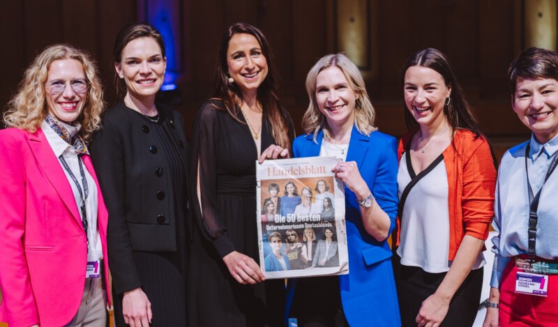 Sechs Frauen lächeln in die Kamera, eine von ihnen hält eine Ausgabe der Zeitung "Handelsblatt", die einen Artikel über die "50 besten Unternehmerinnen Deutschlands" zeigt.