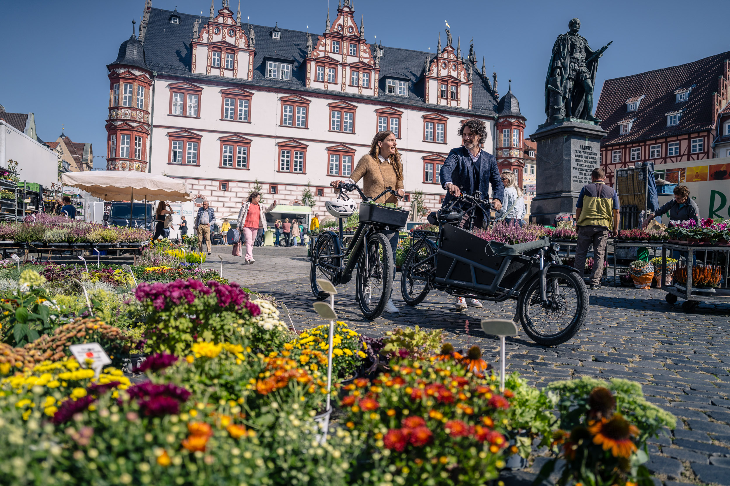 Zwei Personen mit Fahrrädern schlendern über einen bunten Blumenmarkt vor historischer Kulisse.