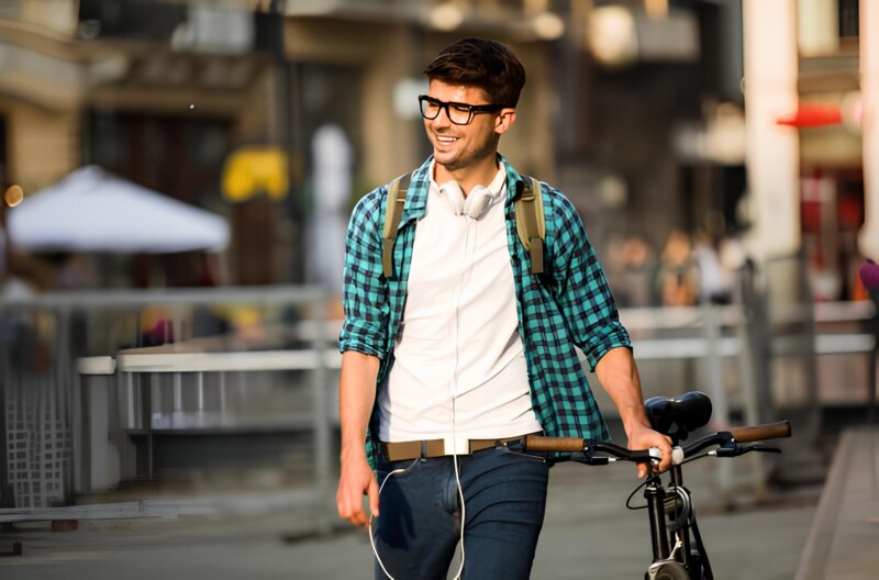 Ein junger Mann mit Brille, Kopfhörern und Rucksack schiebt sein Fahrrad durch eine belebte Stadtstraße und lächelt.