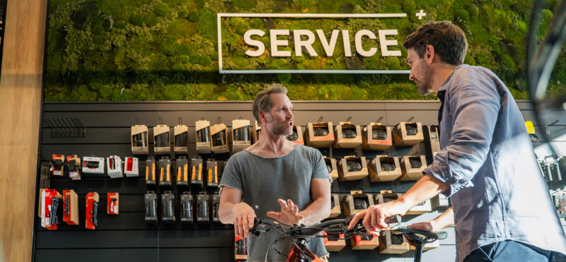 Zwei Männer sprechen in einer Fahrradwerkstatt vor einer Servicewand, während sie sich über ein Fahrrad austauschen.