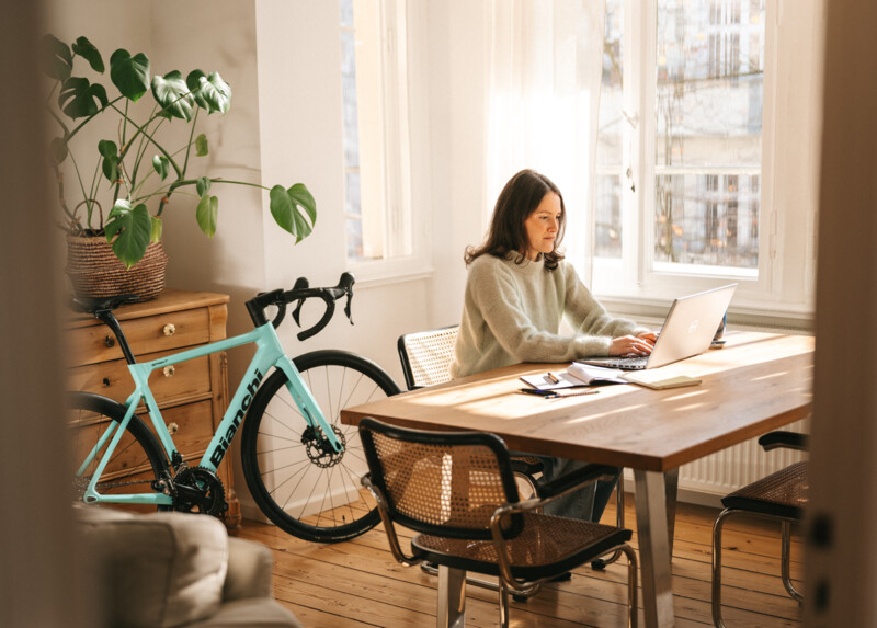 Eine Frau arbeitet an einem Laptop in einem sonnendurchfluteten Raum mit einem Rennrad und einer großen Zimmerpflanze im Hintergrund.