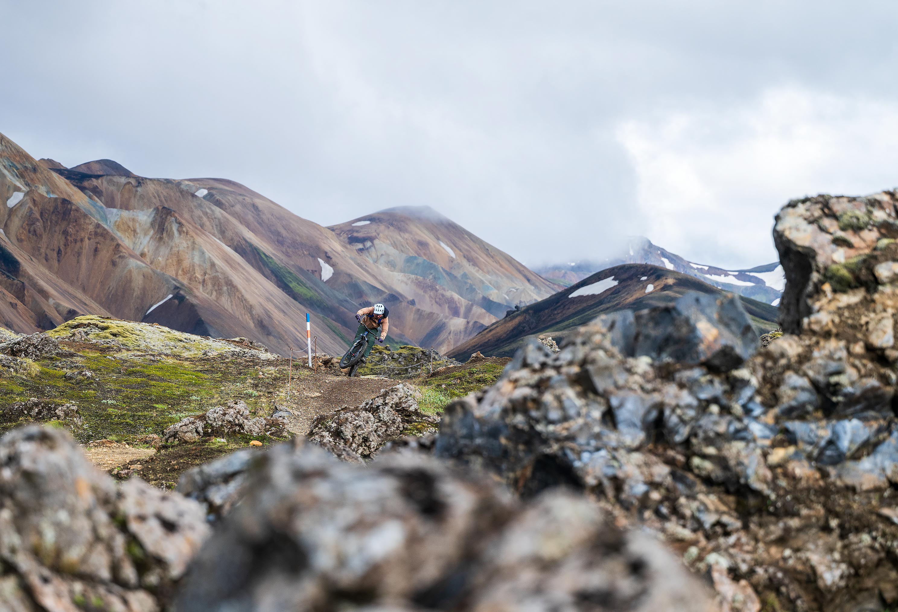 Ein Mountainbiker fährt auf einem schmalen Trail durch eine felsige Landschaft, im Hintergrund farbige Berge mit vereinzelten Schneeflecken.