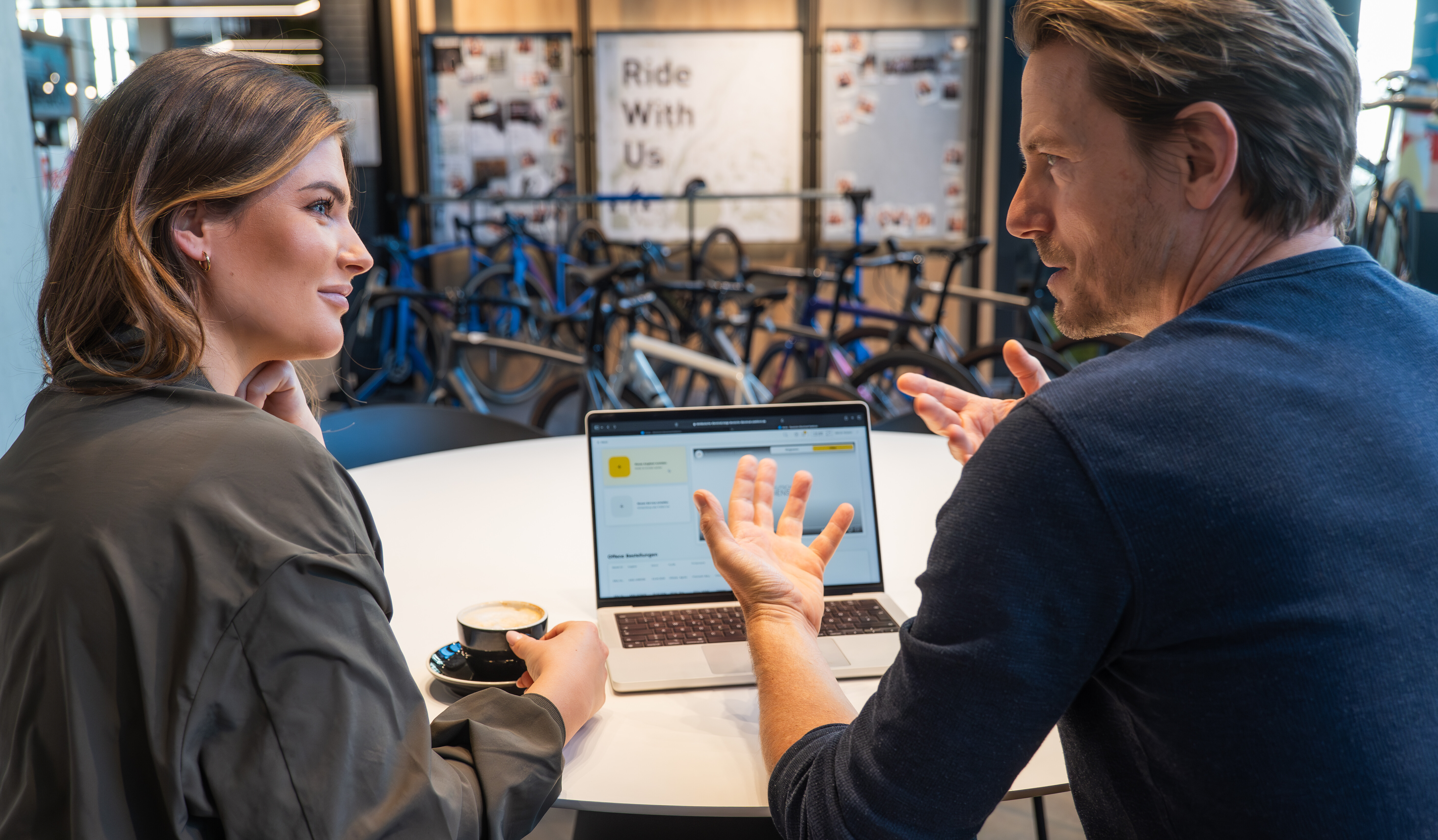 Zwei Personen sitzen an einem Tisch und besprechen etwas am Laptop, während im Hintergrund mehrere Fahrräder in einem Innenraum stehen.