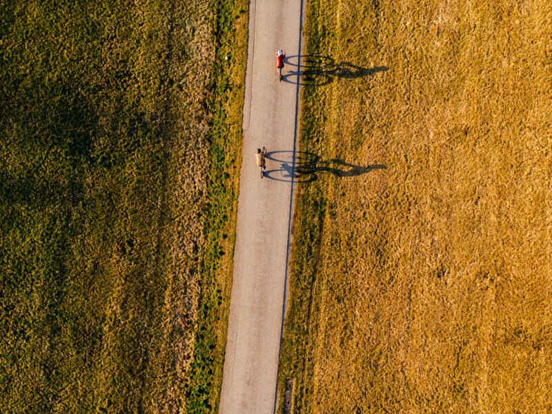 Luftaufnahme eines geraden Radwegs zwischen grüner und goldbrauner Wiese; zwei Radfahrende sind auf dem Weg zu sehen, ihre langen Schatten fallen seitlich auf den Asphalt.