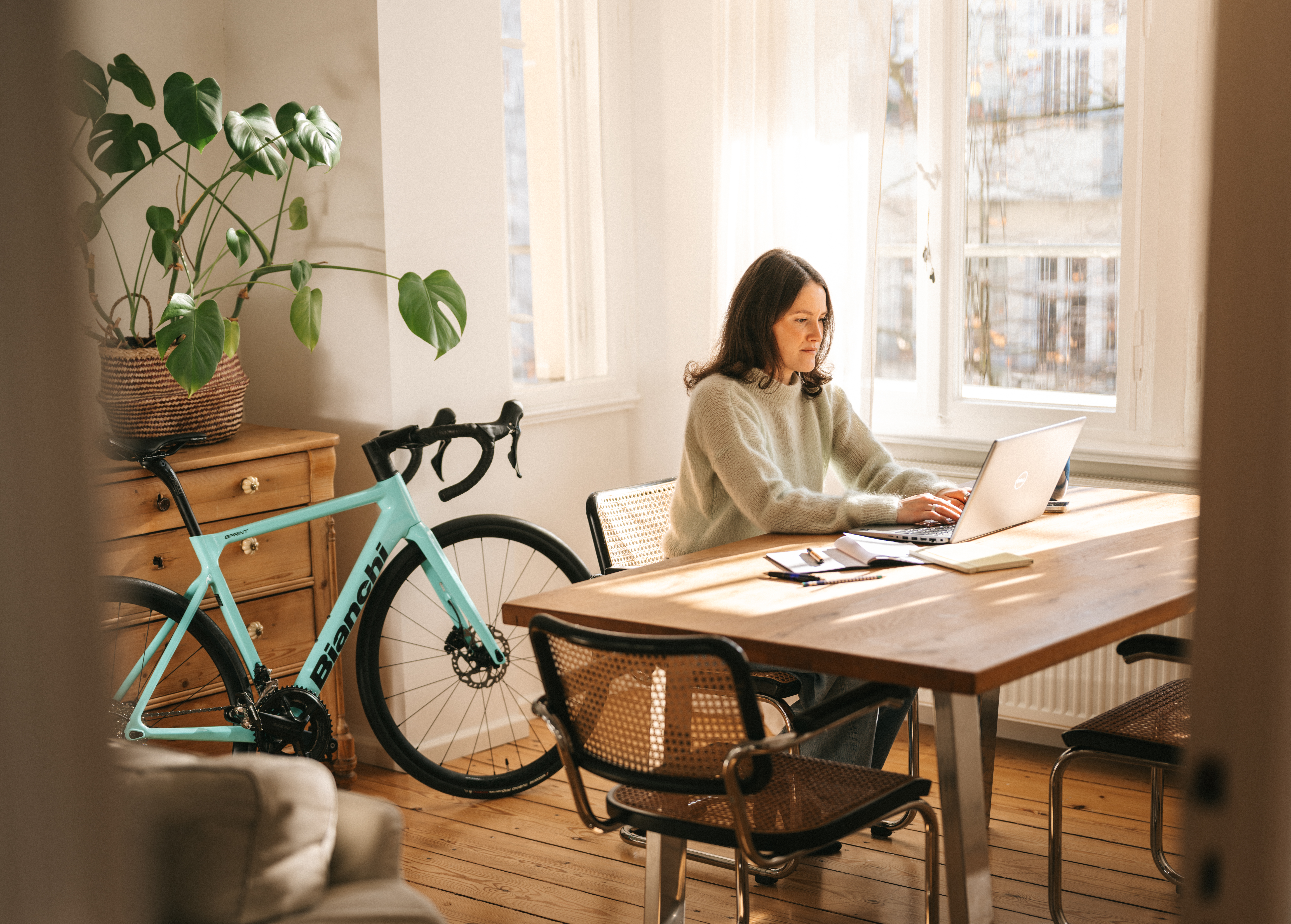 Eine Frau arbeitet an einem Laptop in einem sonnendurchfluteten Raum mit einem türkisfarbenen Rennrad im Hintergrund.